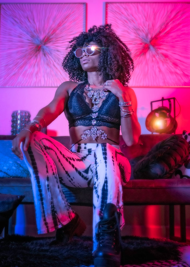 A stylish person with curly hair sits confidently, wearing a black top, patterned pants, and sunglasses, surrounded by colorful lighting.