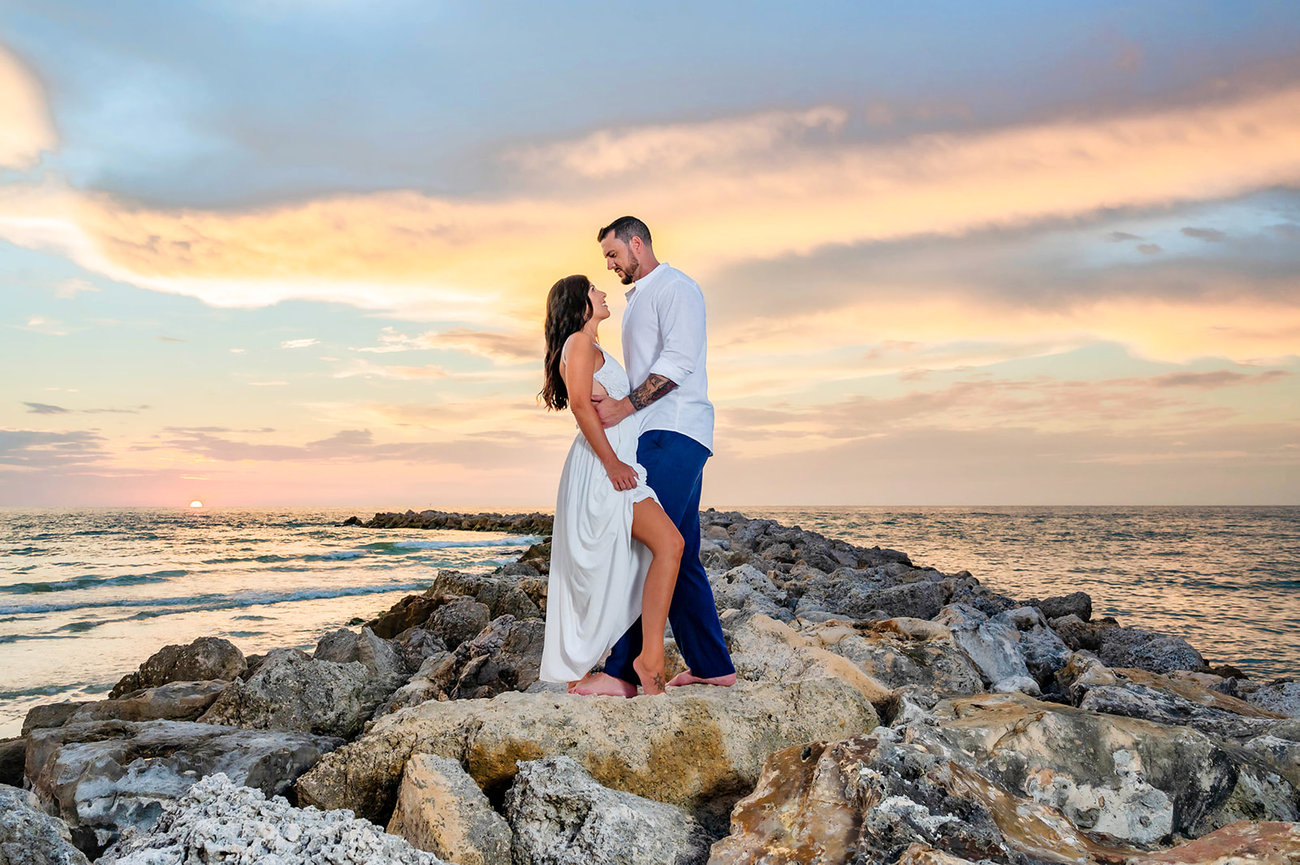 Couple embracing on rocky shoreline at sunset, with vibrant sky and ocean in the background.