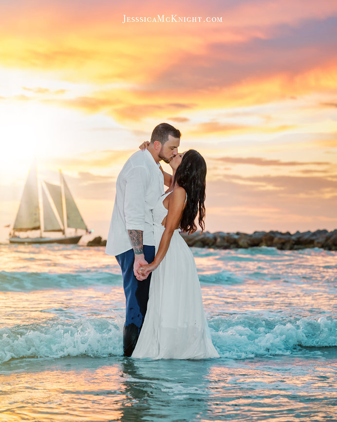 Couple in white attire sharing a kiss in the ocean at sunset, with a sailboat in the background.