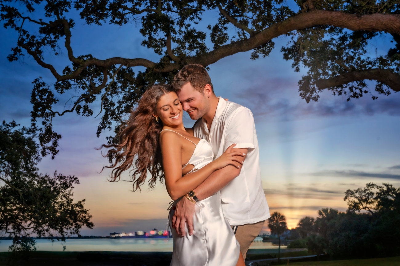 Young couple embracing outdoors at sunset, surrounded by trees and a scenic waterfront backdrop.