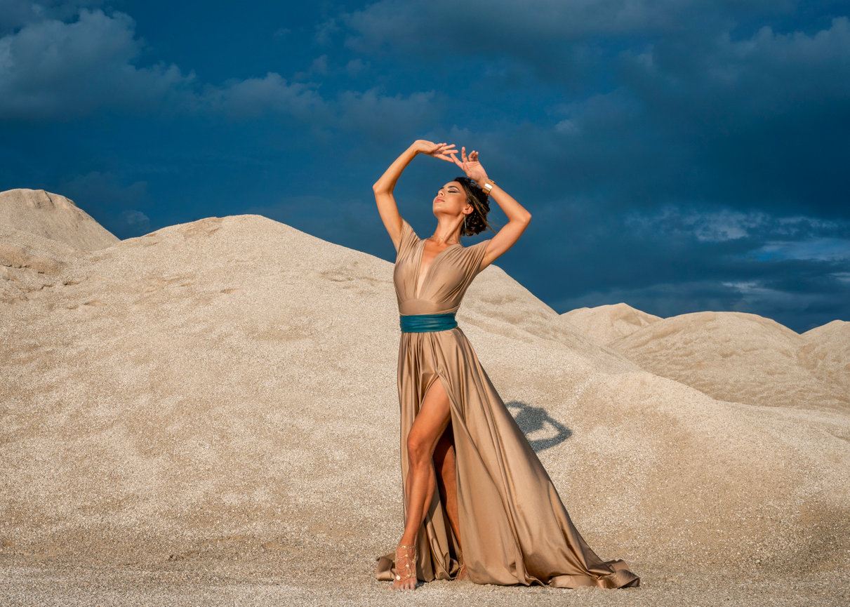 Woman in an elegant bronze gown poses confidently on a sandy hill against a dramatic sky.