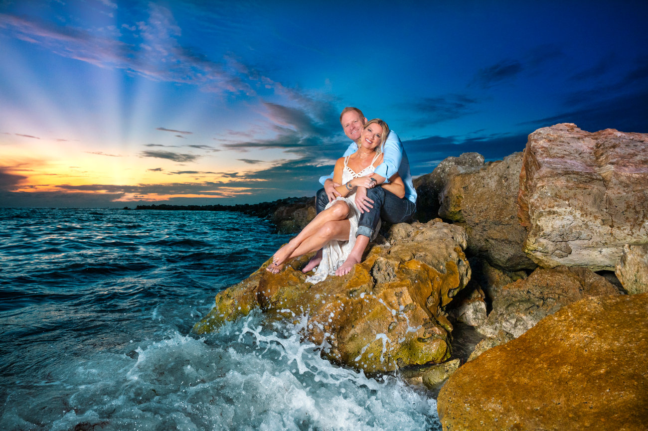 A couple embraces on rocky shoreline at sunset, with waves crashing and a colorful sky in the background.