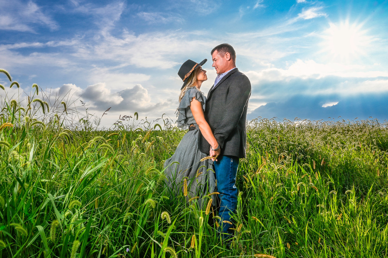 Couple holding hands in a field at sunset, wearing casual and vintage attire, with bright blue sky and clouds.