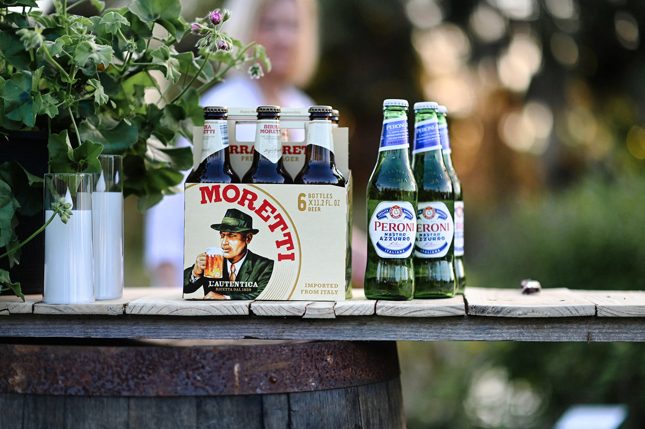 A selection of Moretti beer and Peroni bottles displayed on a wooden barrel with greenery in the background.