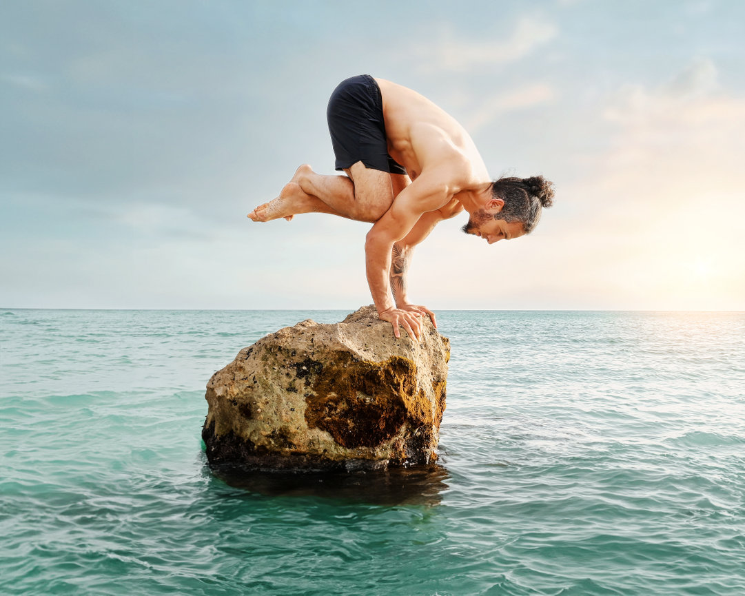 Fit man performing a yoga pose on a rock above water at sunset.