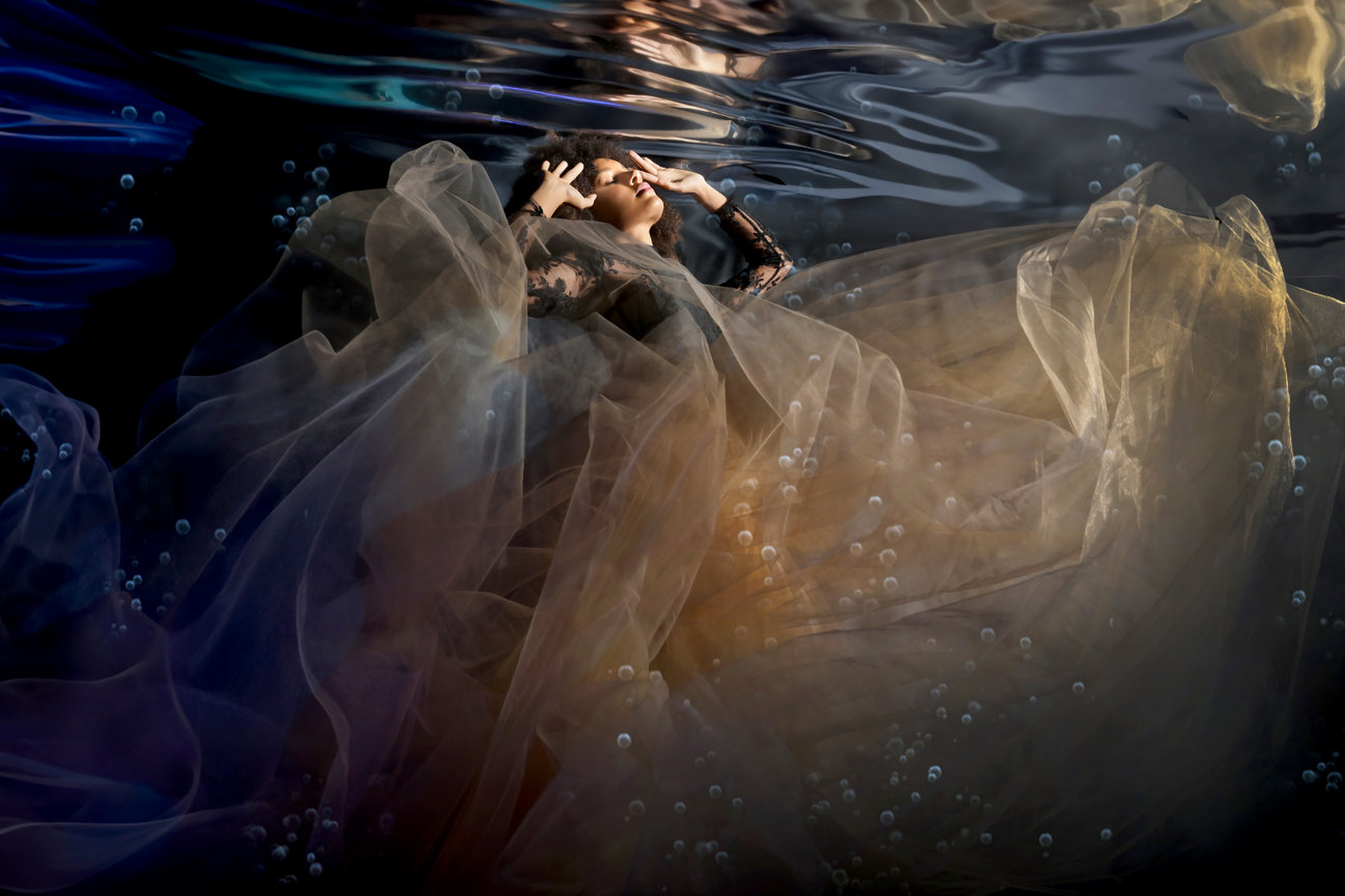 A woman in a flowing gown floats underwater, surrounded by bubbles and soft fabric, creating a serene, ethereal atmosphere.