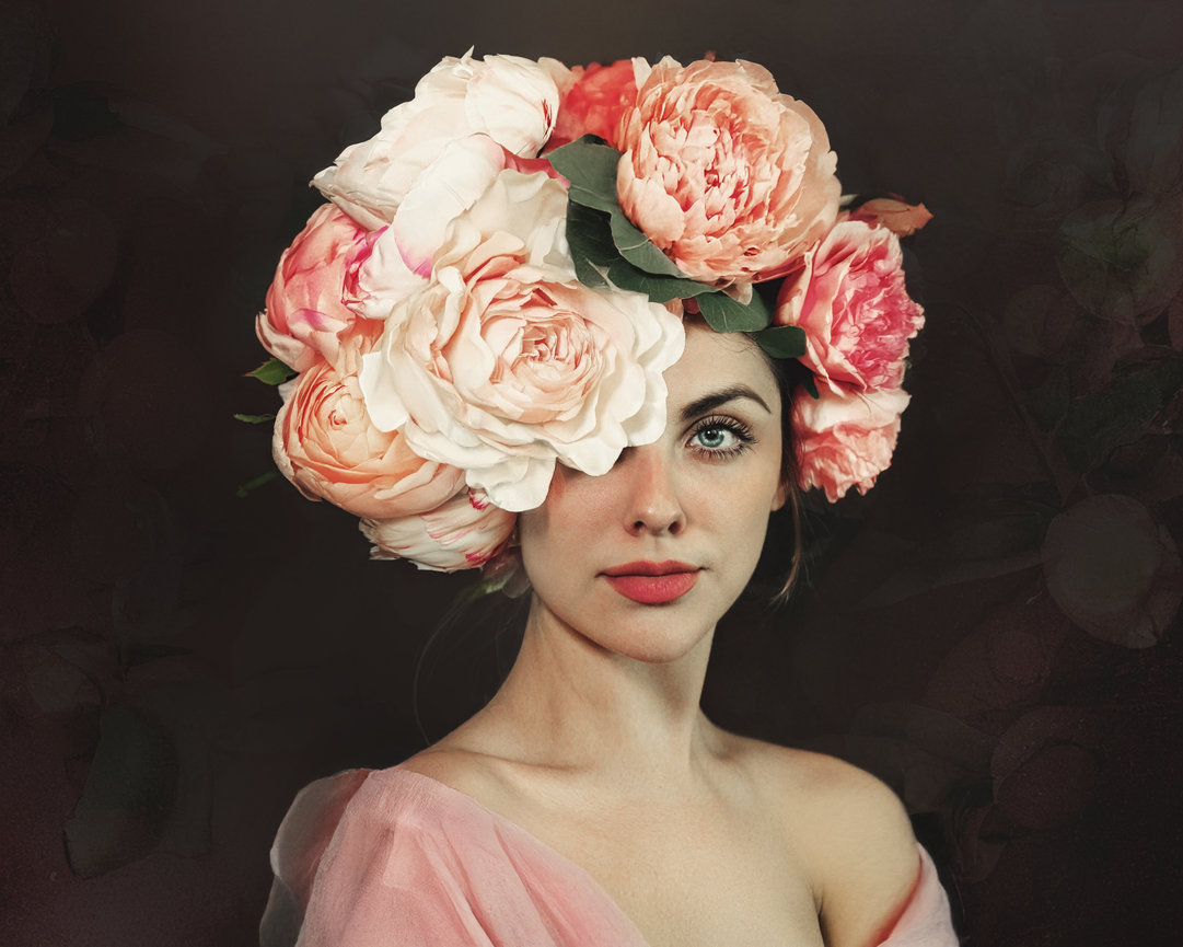 Woman with floral crown made of pink and cream peonies, wearing a soft pink garment and a natural expression.