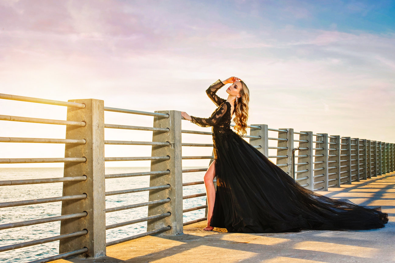 Woman in a long black gown poses on a seaside pier, with sunlight reflecting on the water behind her.