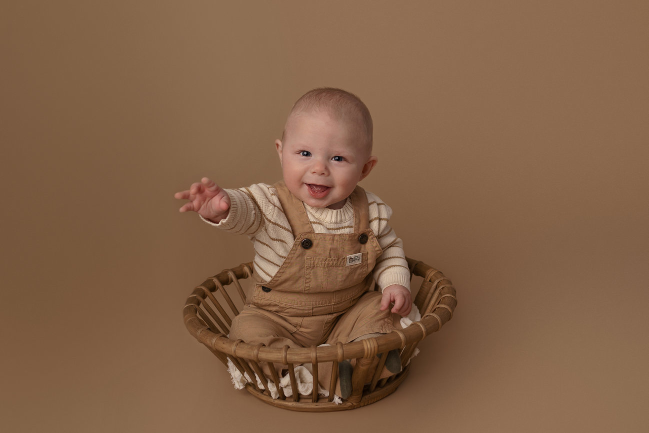 Smiling baby in striped shirt and overalls sitting in a round basket on a brown background.