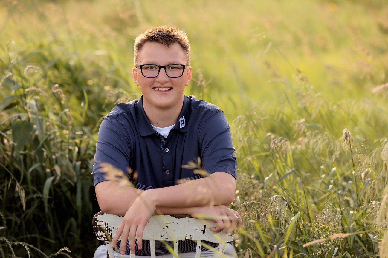 Person sitting on a chair in a grassy field, wearing glasses and a blue shirt, smiling at the camera.