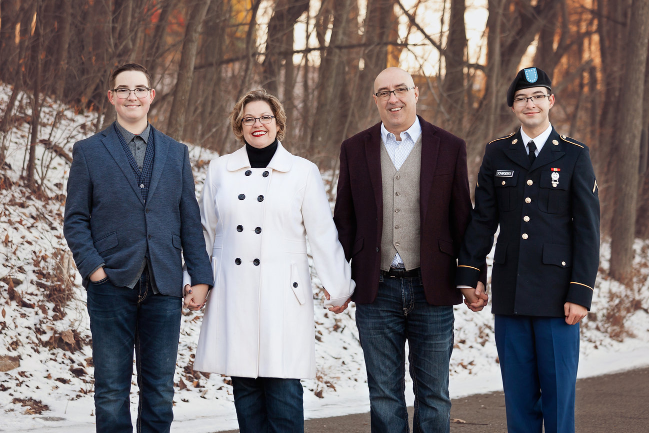Family of four holding hands, standing outdoors on a snowy path. One in military uniform, others in winter clothing.