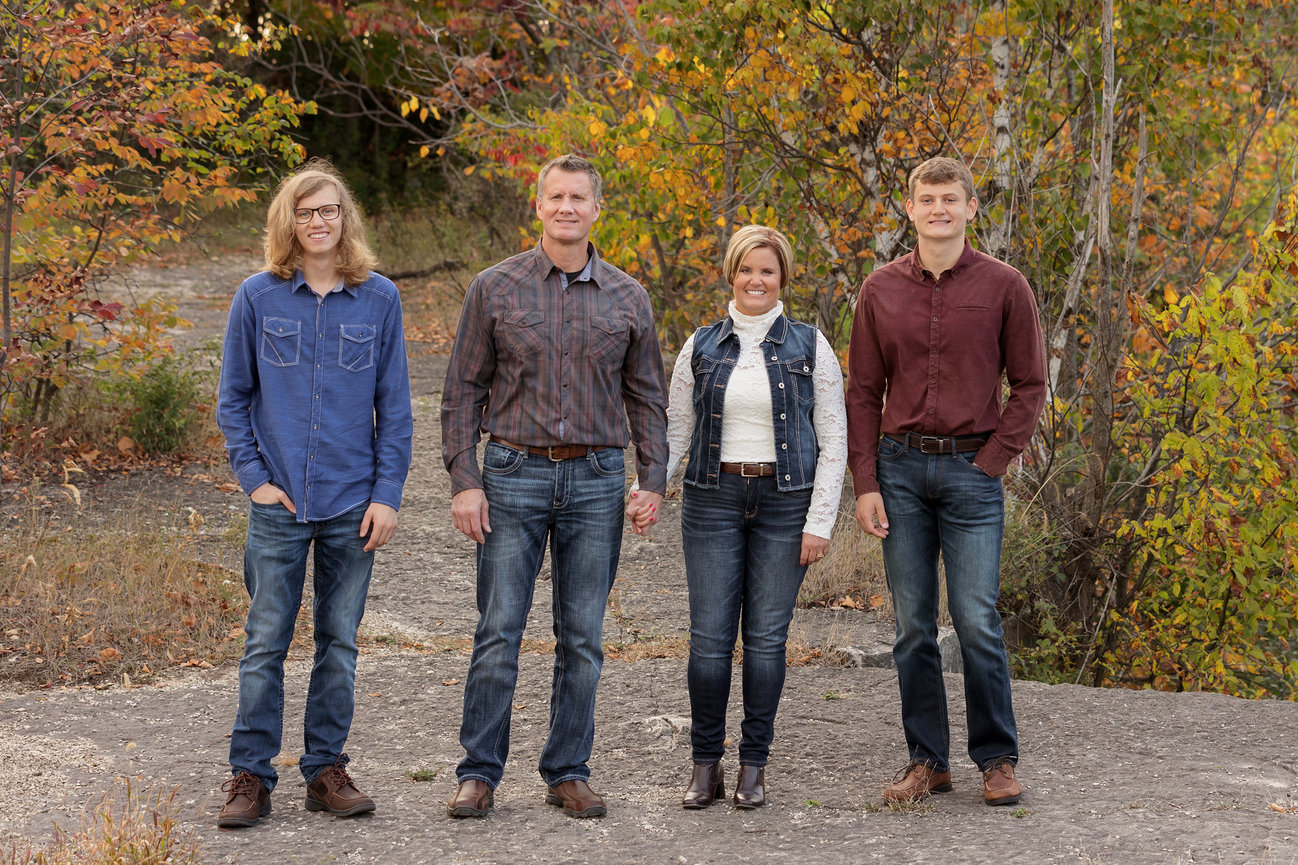 Family of four standing outdoors on a rocky path surrounded by autumn foliage, dressed in casual clothing.