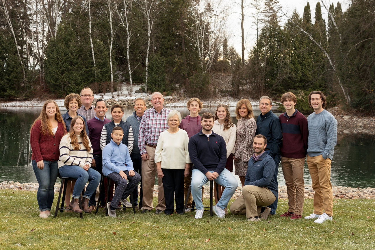 A large family poses together outdoors by a lake, with evergreen trees in the background. They are dressed in winter clothing.