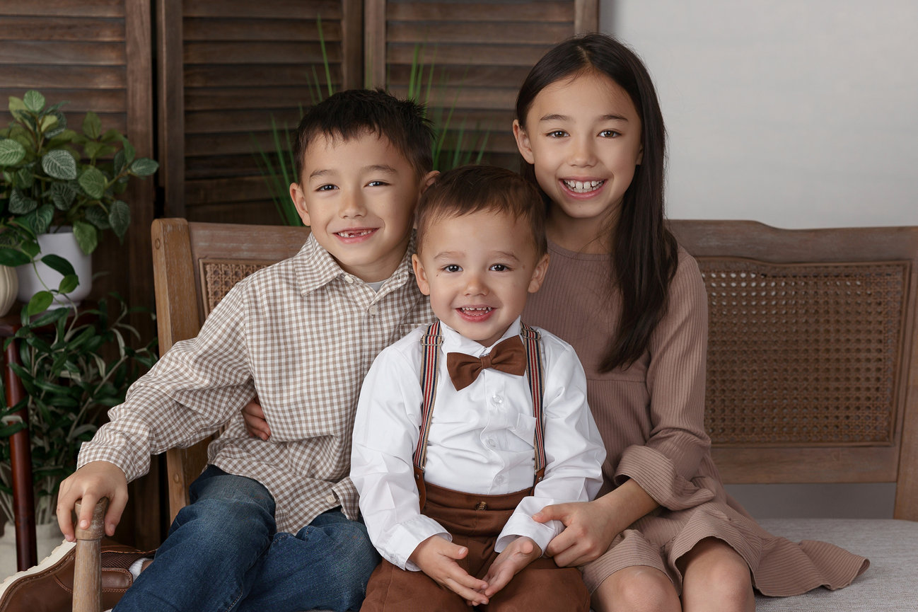 Three smiling children seated on a wooden bench, plants in the background, dressed in coordinated outfits.