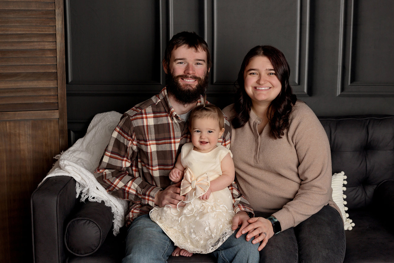 A smiling couple sits on a couch with a baby in a white dress between them.