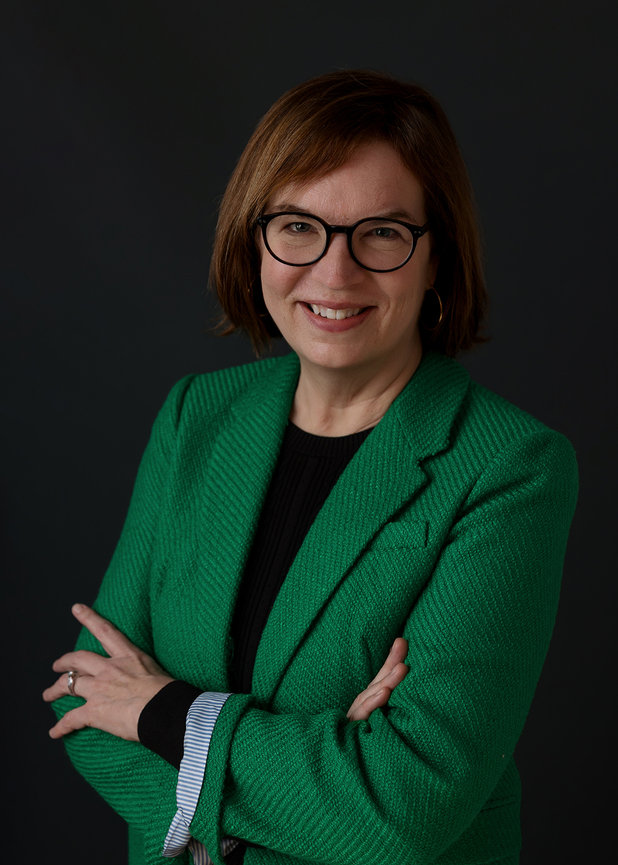 Person with glasses and brown hair wearing a green blazer, smiling with arms crossed against a dark background.