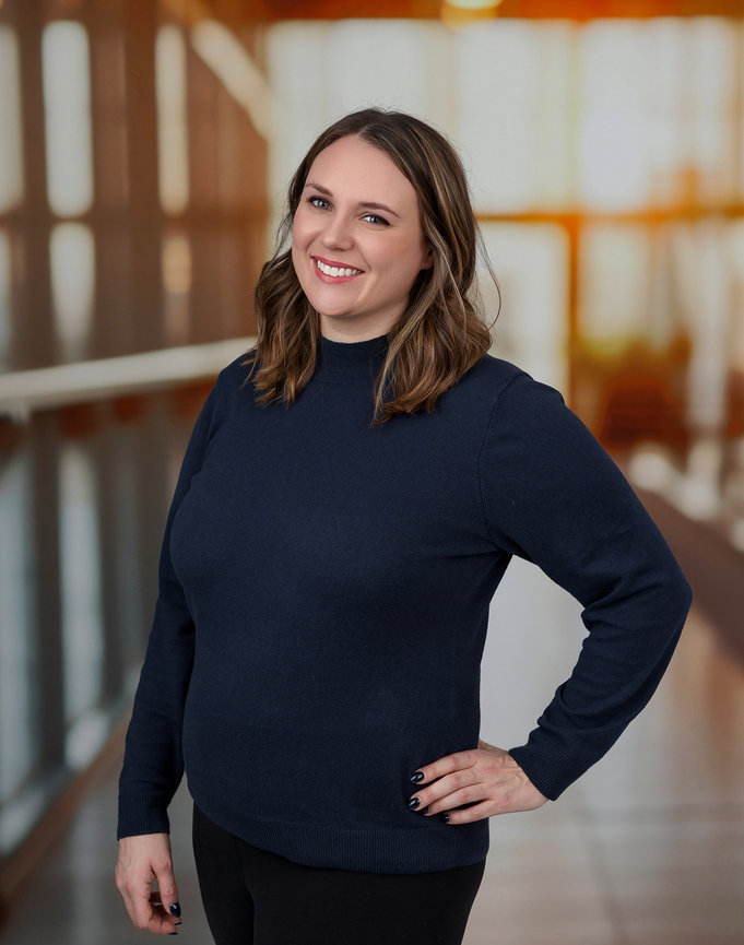 Smiling person with brown hair in a blue sweater stands in a sunlit indoor space.