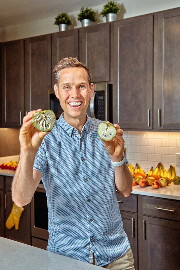 Man smiling, holding two halves of a cucumber in a kitchen. Dark cabinets and fruit visible in the background.