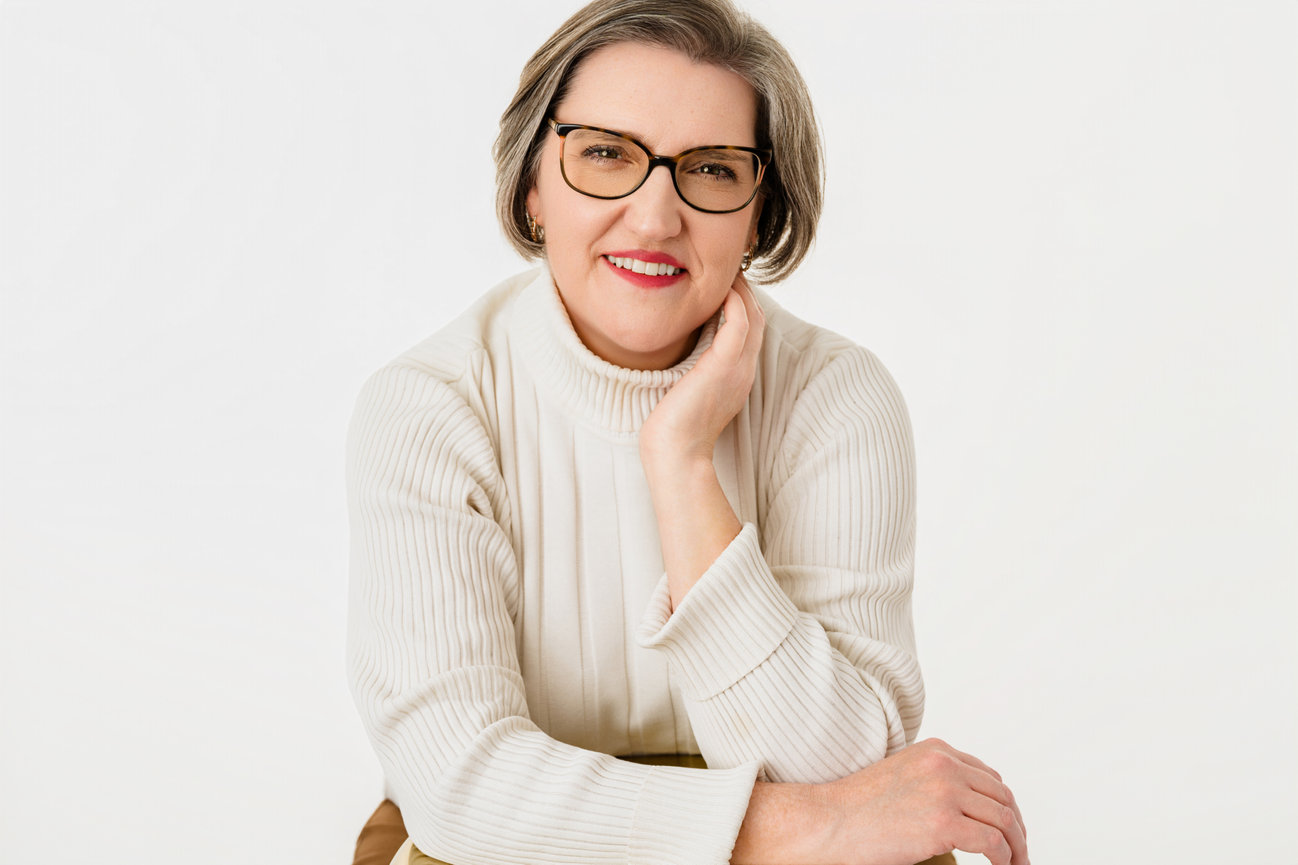 Woman in glasses and white sweater smiles, resting head on hand against a white background.