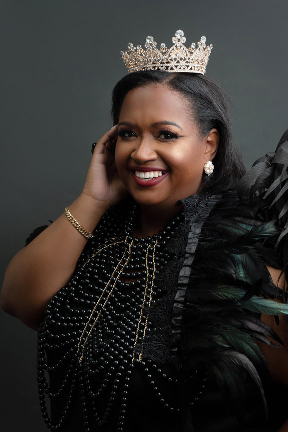 Smiling woman wearing a crown and black pearls, dress, and angel wings, hand on face.