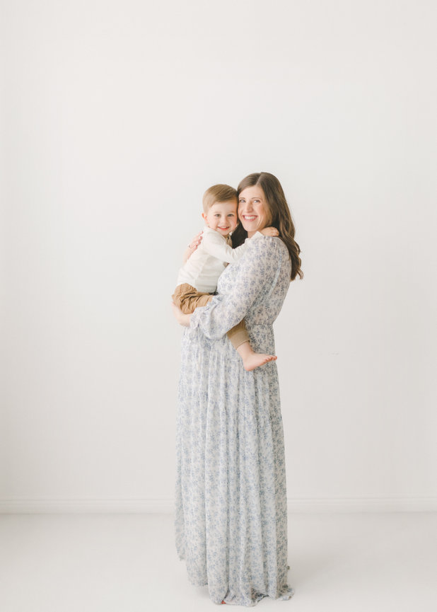 A woman in a floral dress holds a smiling child against a light backdrop.