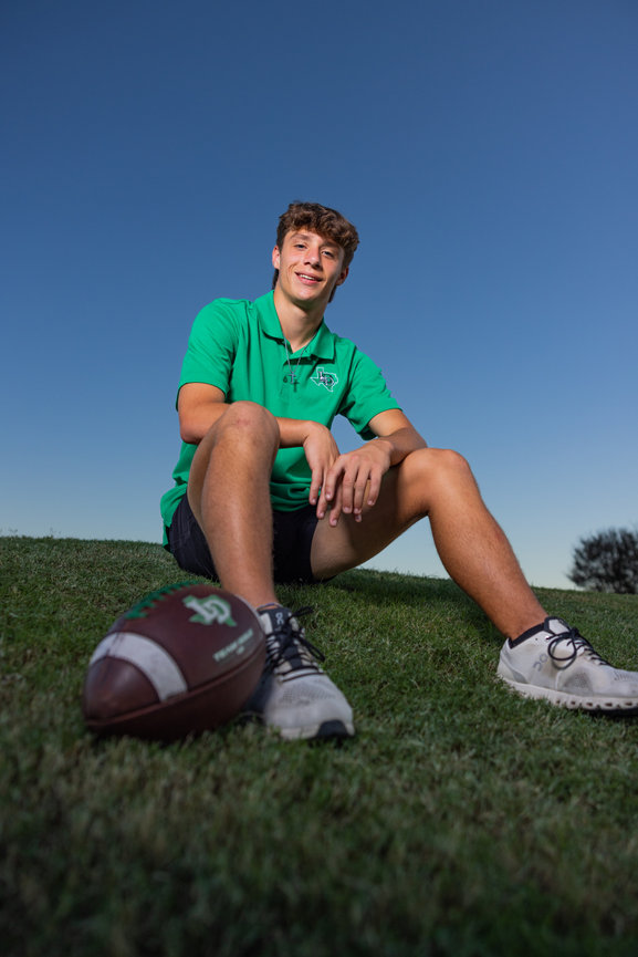 High school senior guy wearing green polo shirt sitting on a green grass hill with a football.