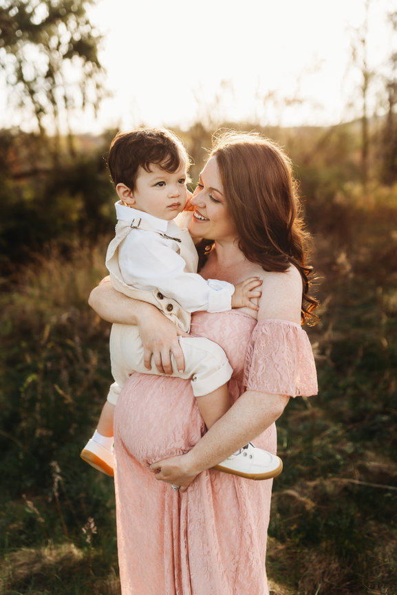 An expecting mother in a pink dress holds her son during sunset at a New Jersey maternity photography session.