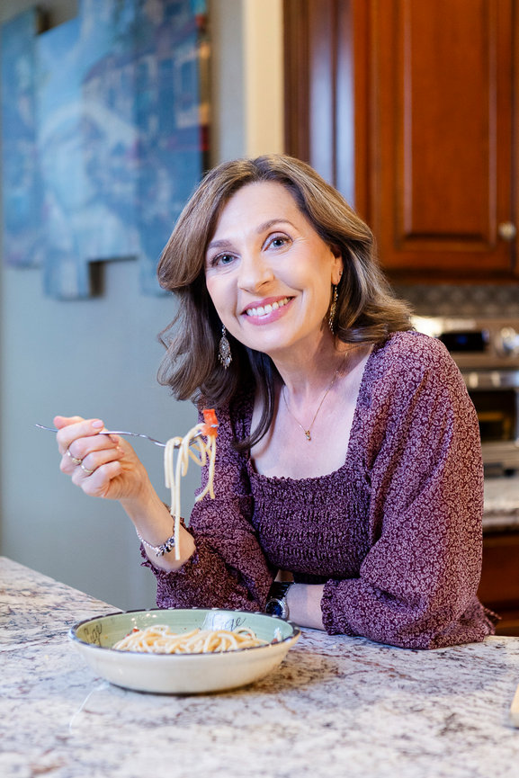 Woman smiling while eating spaghetti at a kitchen countertop.