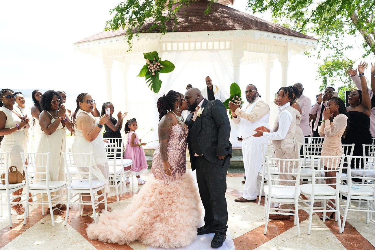 A couple kisses in front of a gazebo, surrounded by clapping guests in formal attire.