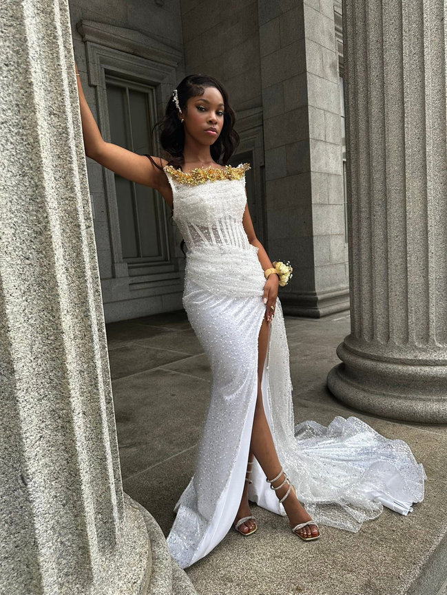 Woman in an elegant white gown with gold accents stands by stone columns, showcasing a high slit and flowing train.