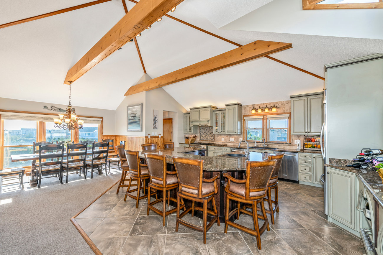 Spacious kitchen and dining area with wooden beams, large island, chairs, and ample natural light through large windows.
