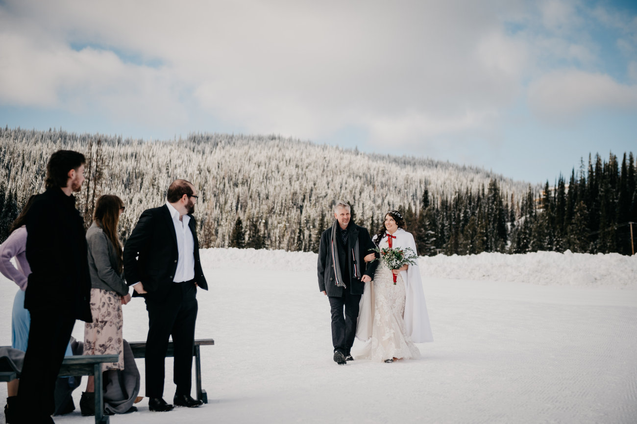 Bride walking aisle: Bride walks down the aisle with her father, framed by mountain snow and panoramic Okanagan views.