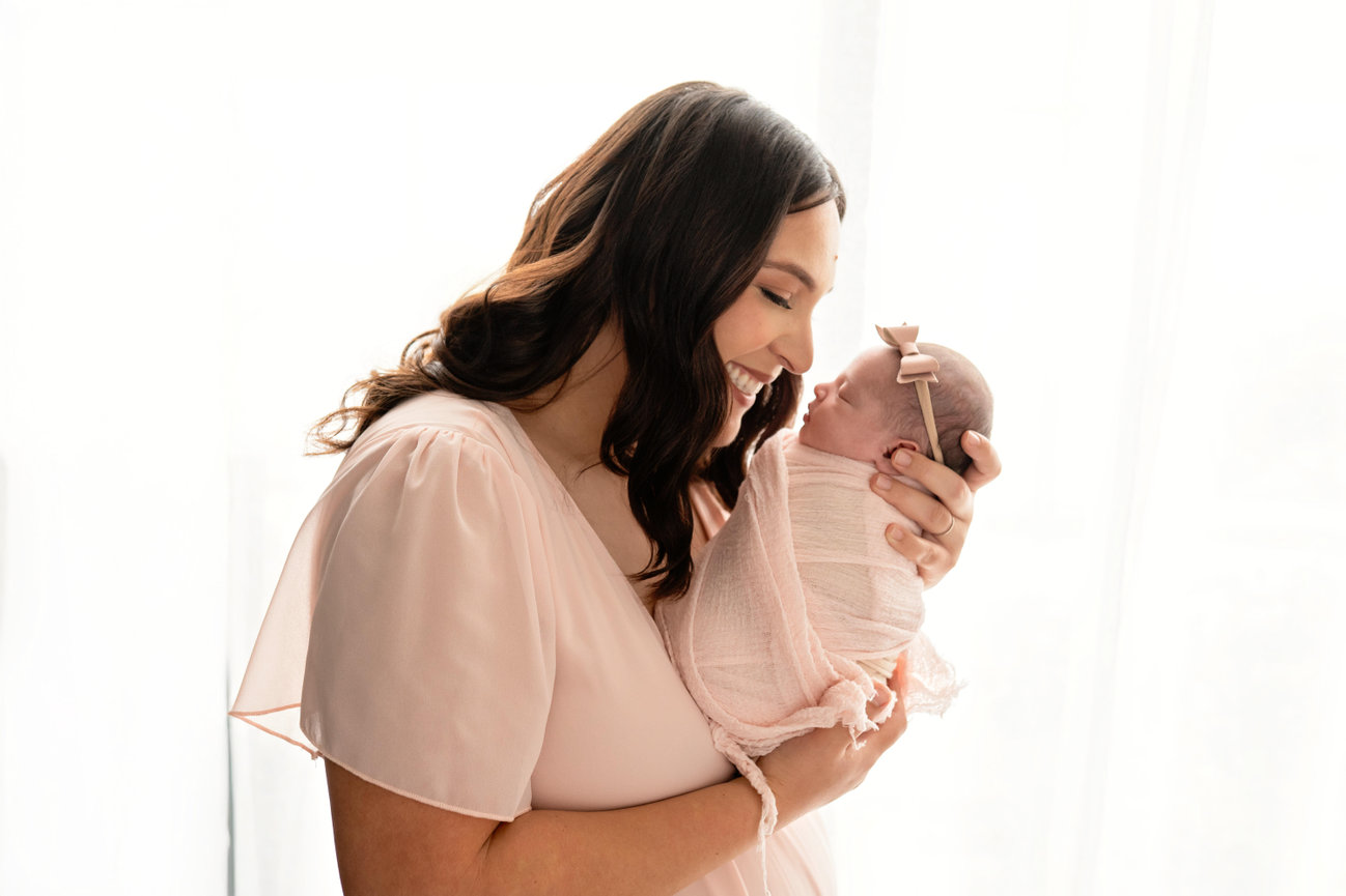 Smiling mother gently holding newborn baby in pink blanket and headband against a bright white background