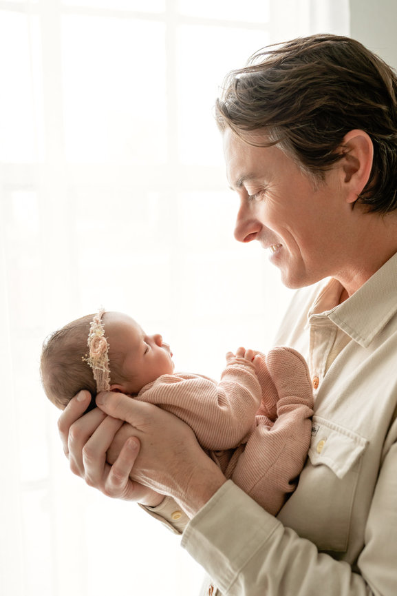 Man holding a sleeping baby girl in soft pink clothes by a bright window