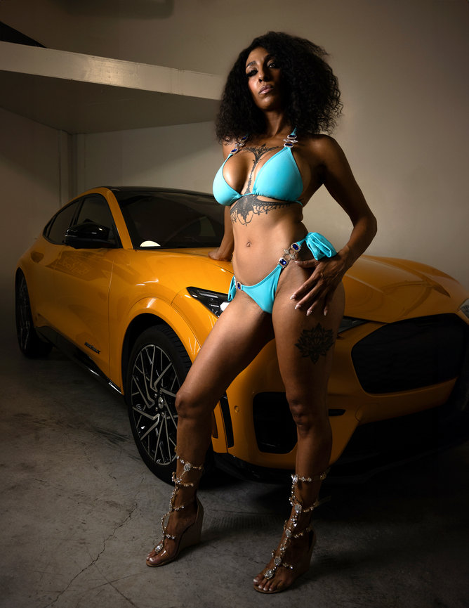 African-American model in a blue bikini posing confidently in front of a yellow Ford Mustang Mach-E GT in a garage setting in Bountiful, Utah. Captured by Tre Nelson Photo, blending bold fashion with automotive elegance.