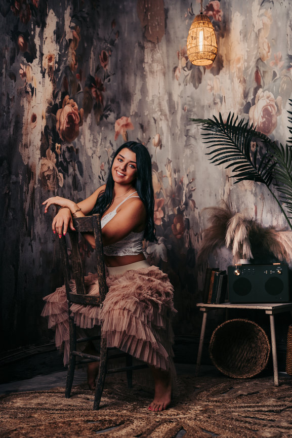 Senior girl posing on a chair in a floral room with a woven lamp and tropical decor