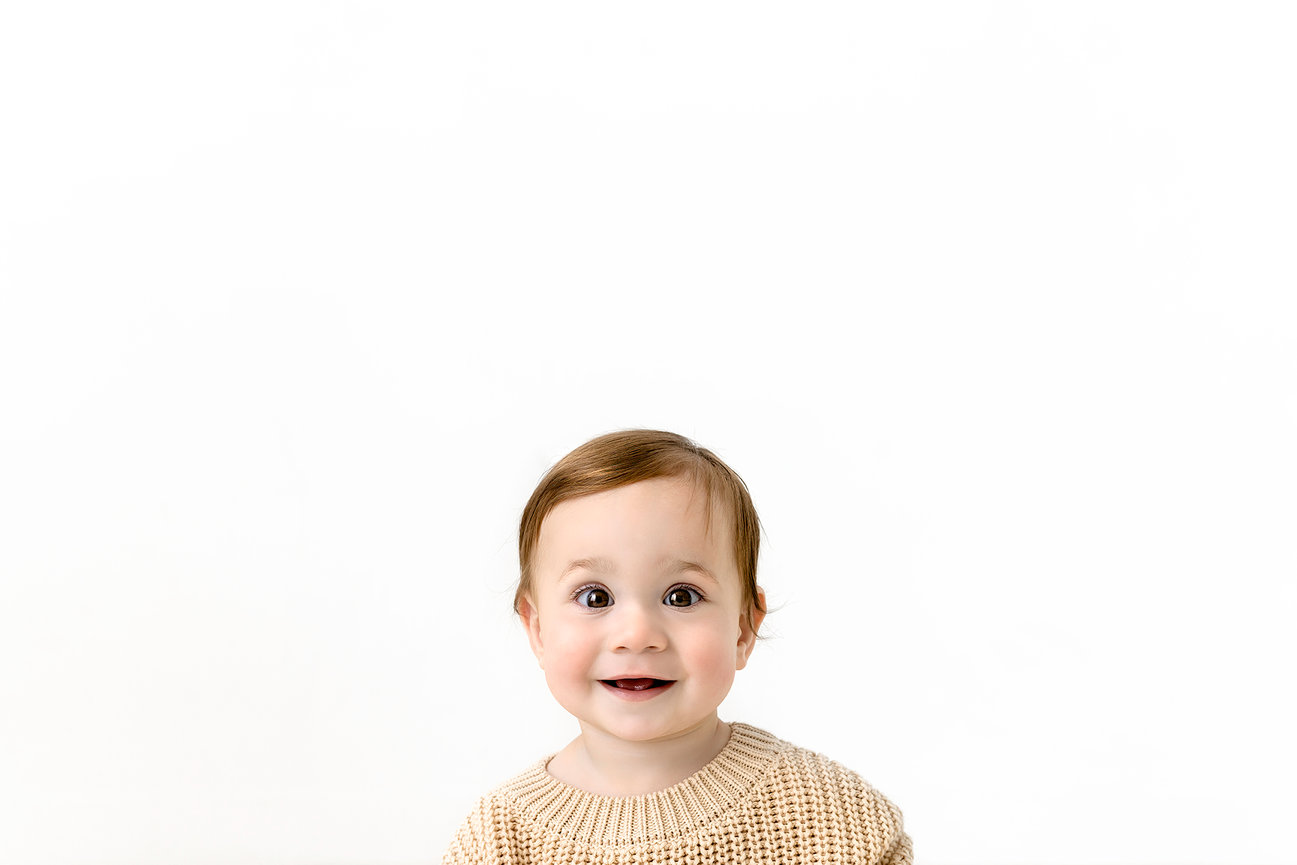 Smiling baby with auburn hair in a beige sweater on a white background.