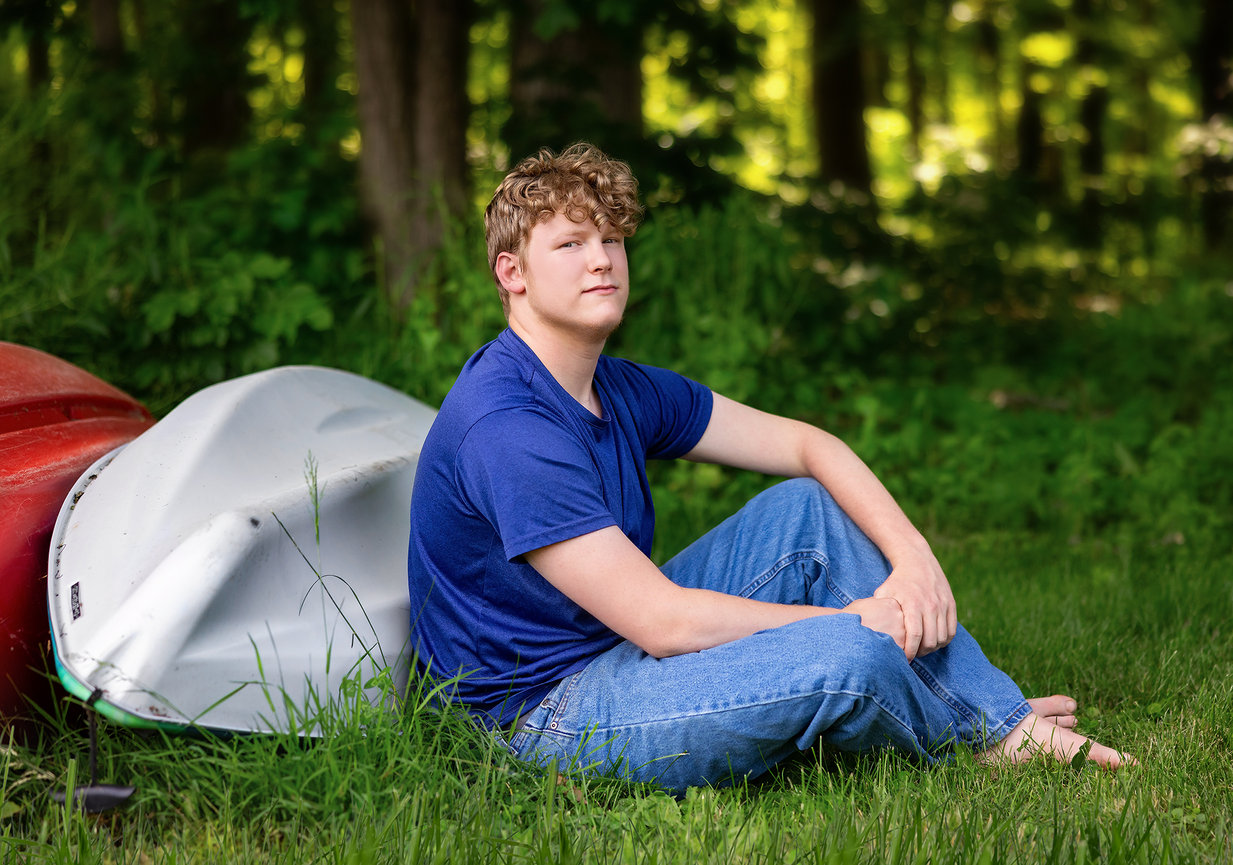 Teen sitting on grass beside overturned kayaks in a wooded area