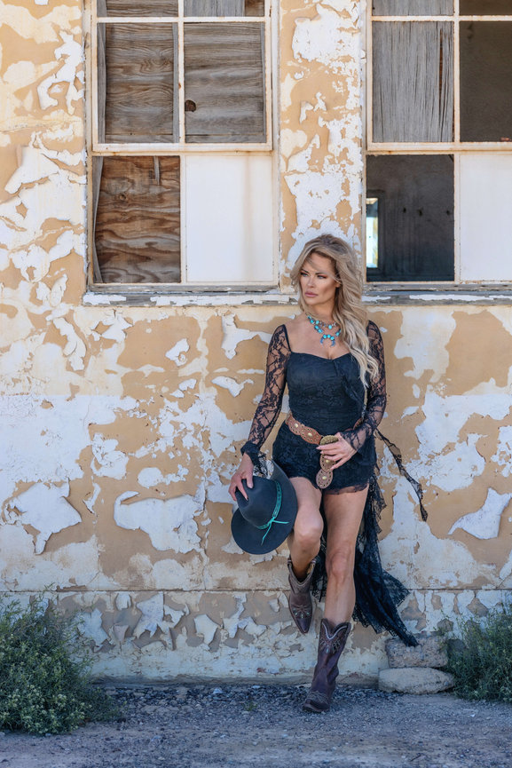Woman in black lace outfit and cowboy boots posing against a weathered wall with boarded windows