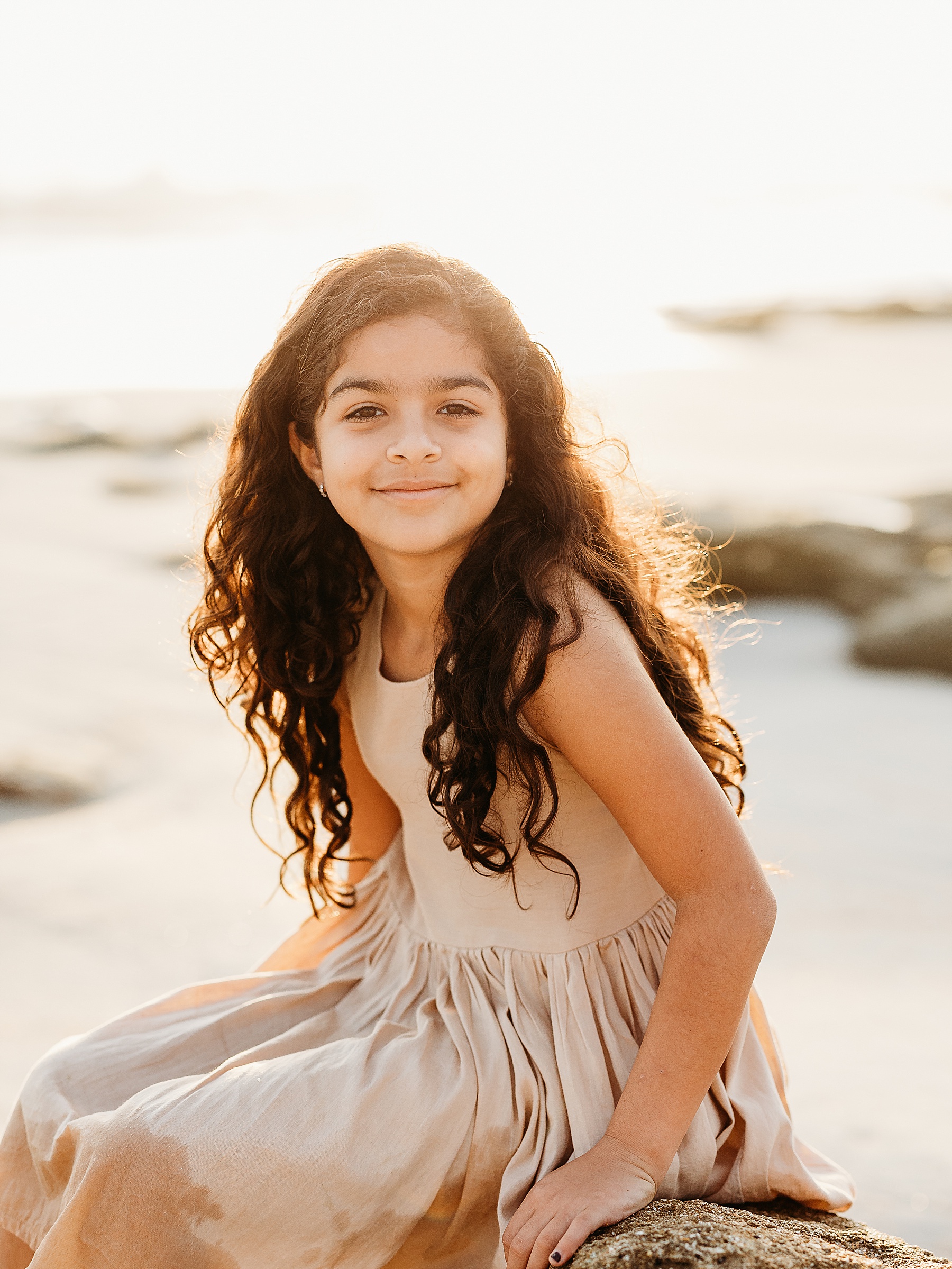 little girl sitting on the rocks at the beach wearing a nude colored dress