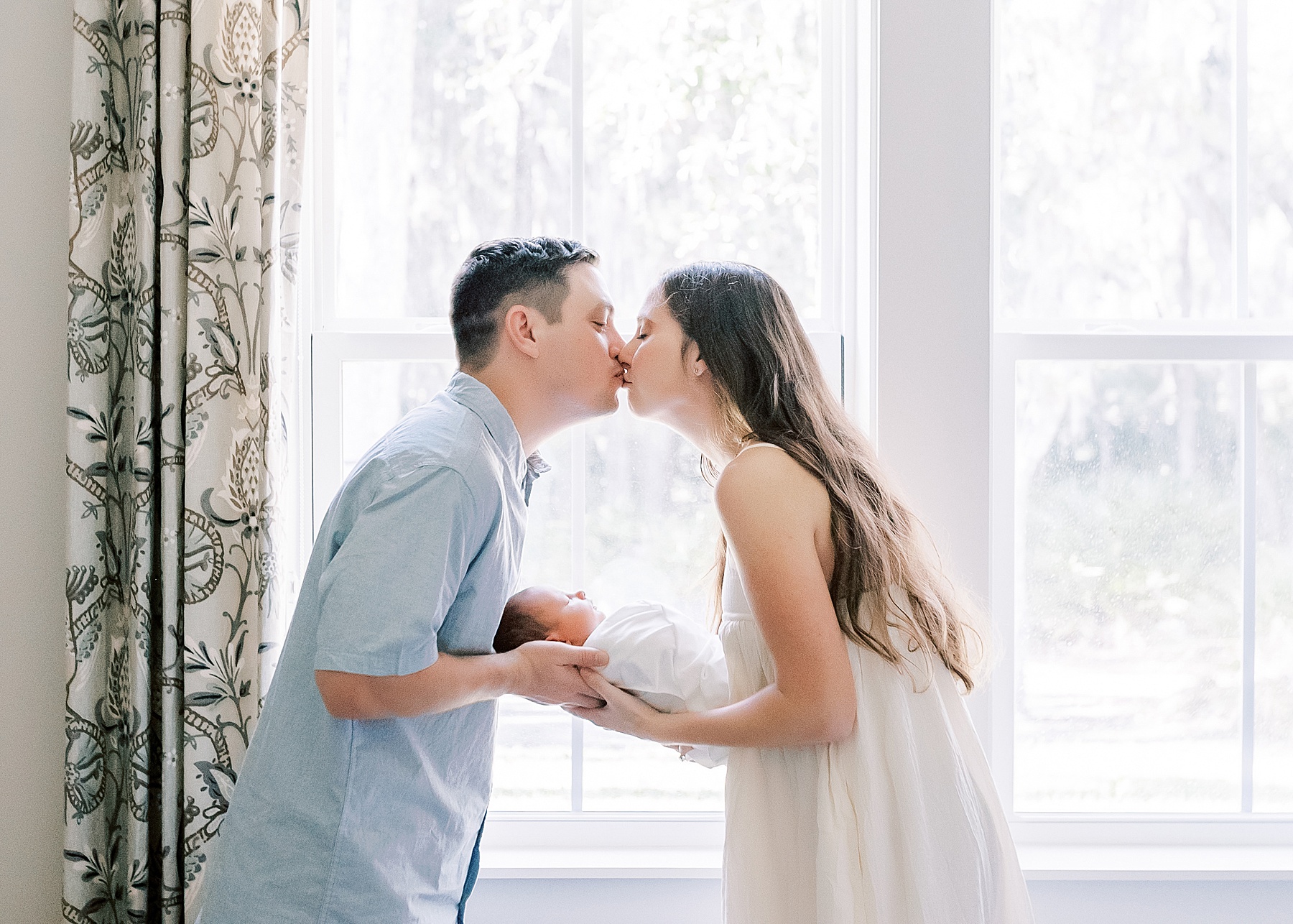 man and woman kissing in front of window while holding newborn baby boy