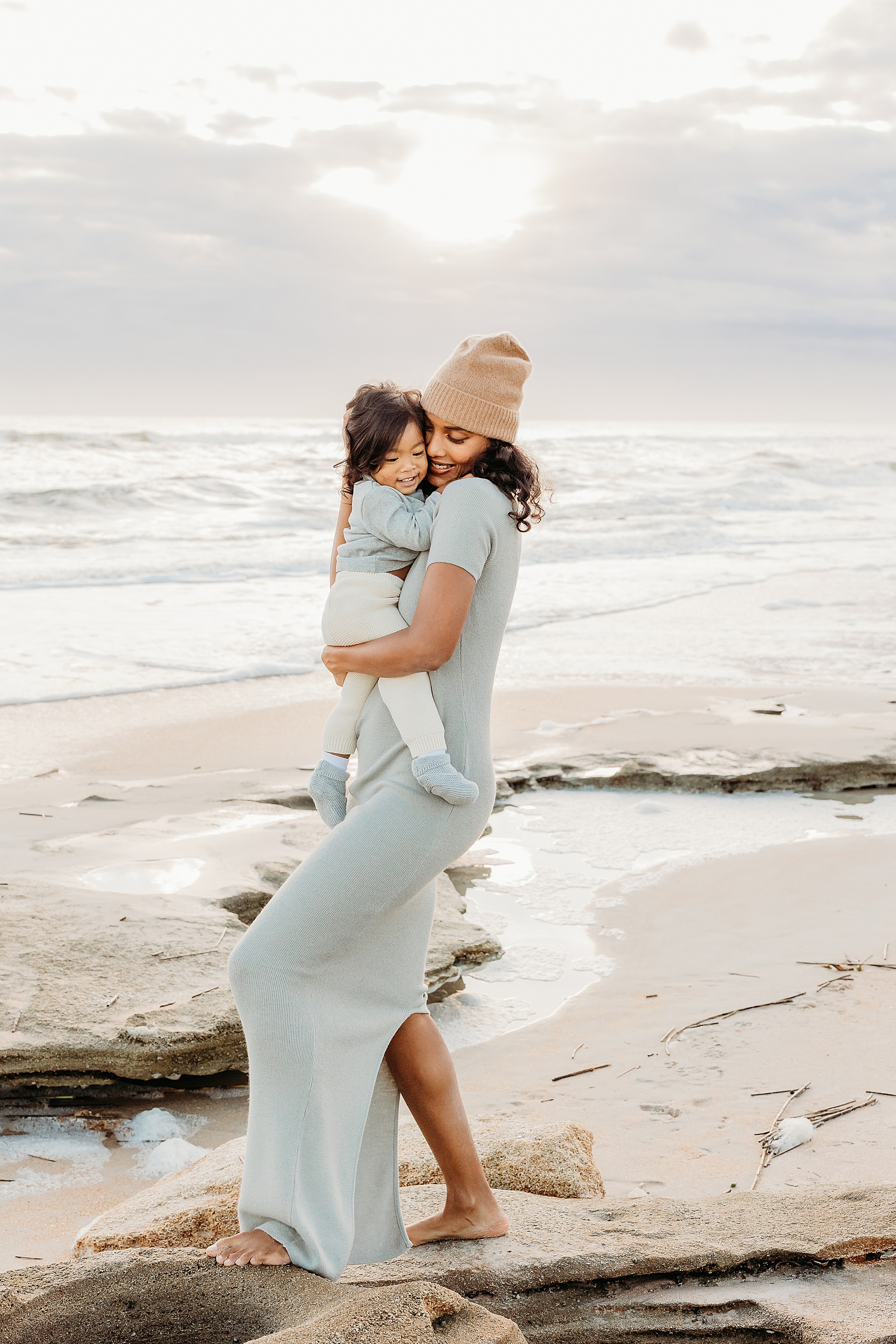 woman wearing green maxi dress holding baby boy on the beach at sunrise smiling