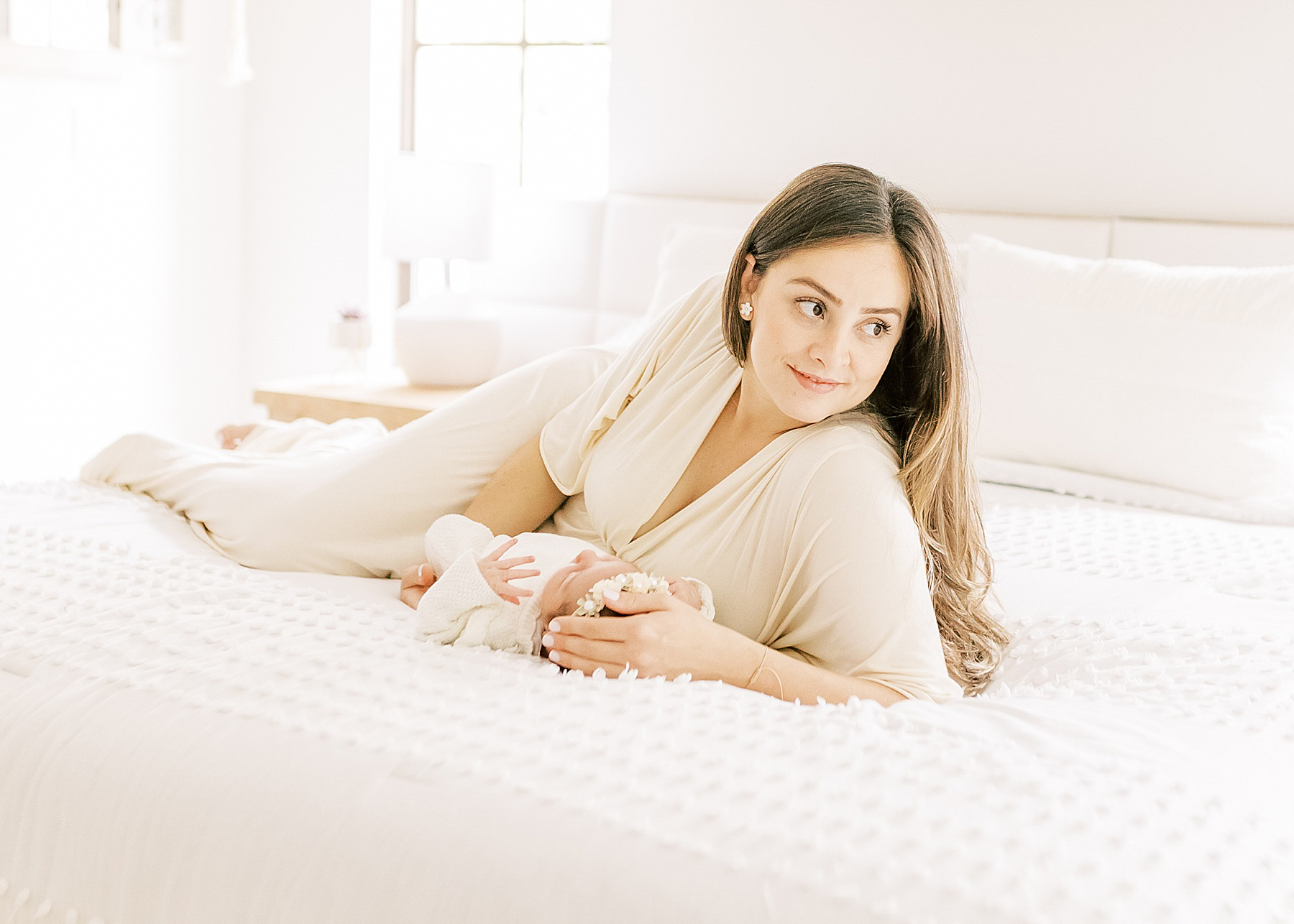 woman with long dark hair in cream long dress sitting on bed smiling with baby