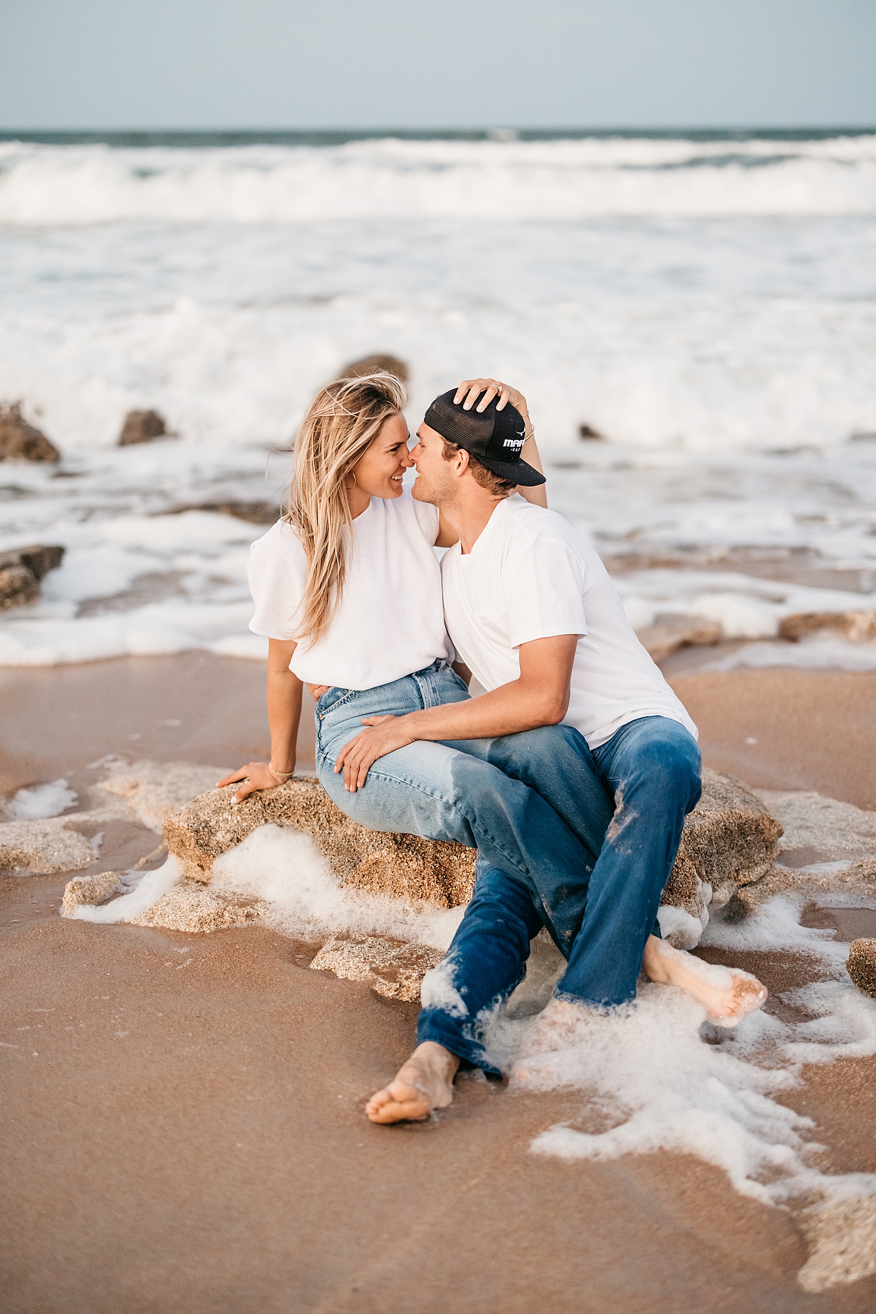 woman in white shirt and jeans sitting on rocks at the beach with man in jeans