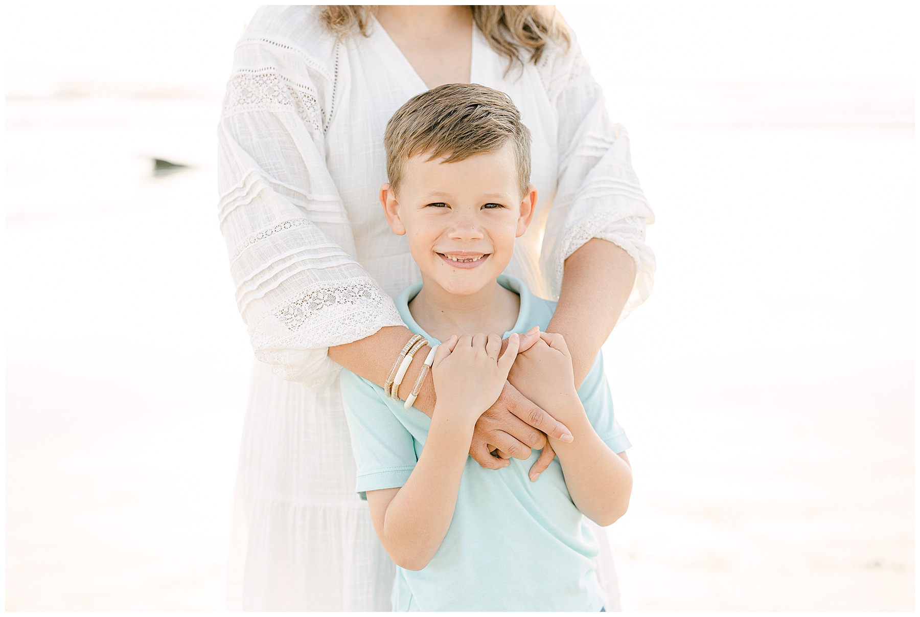 little boy holding onto mom smiling on the beach at sunrise