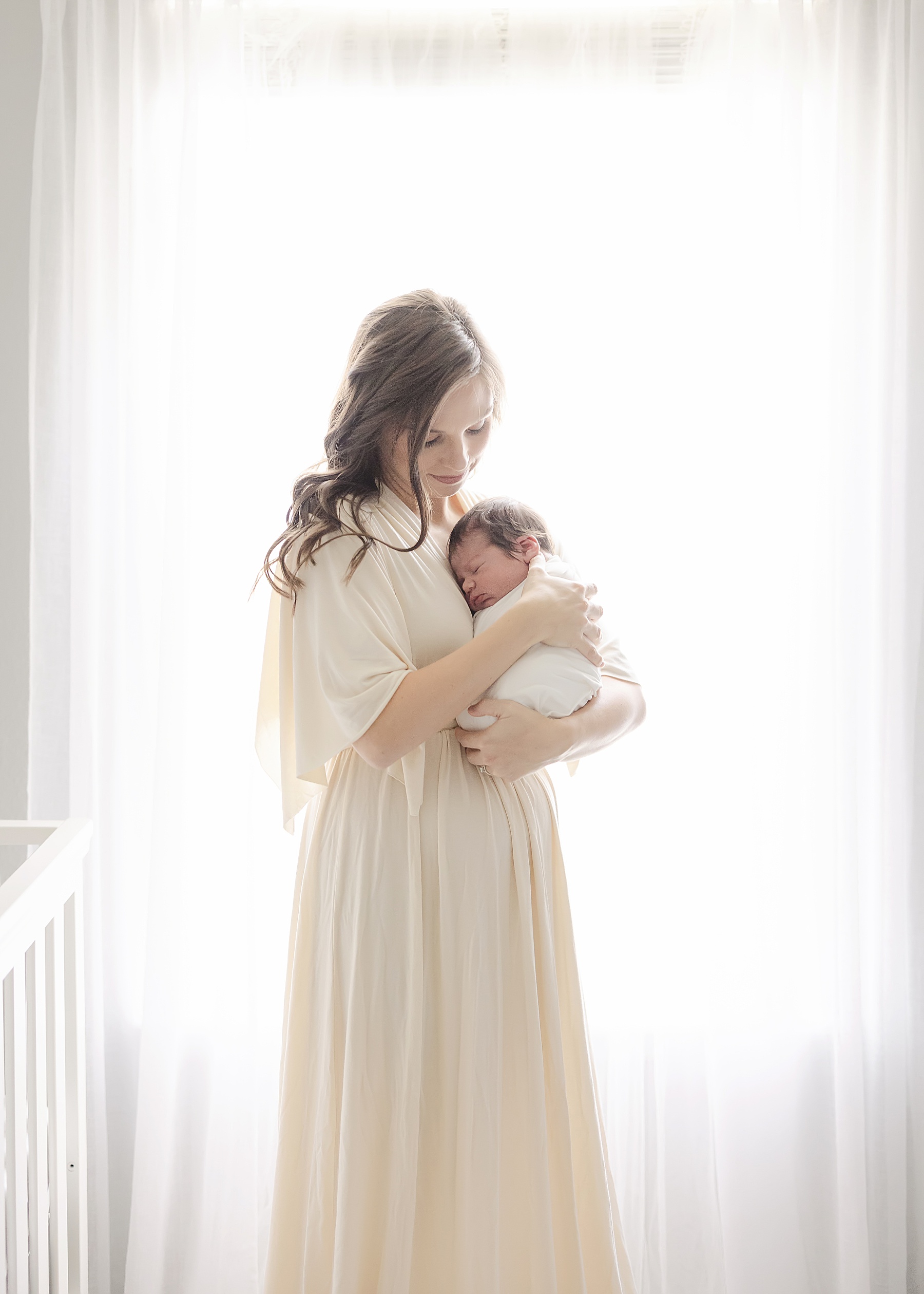 woman wearing long cream dress during in home newborn session holding newborn baby boy by window with white sheers