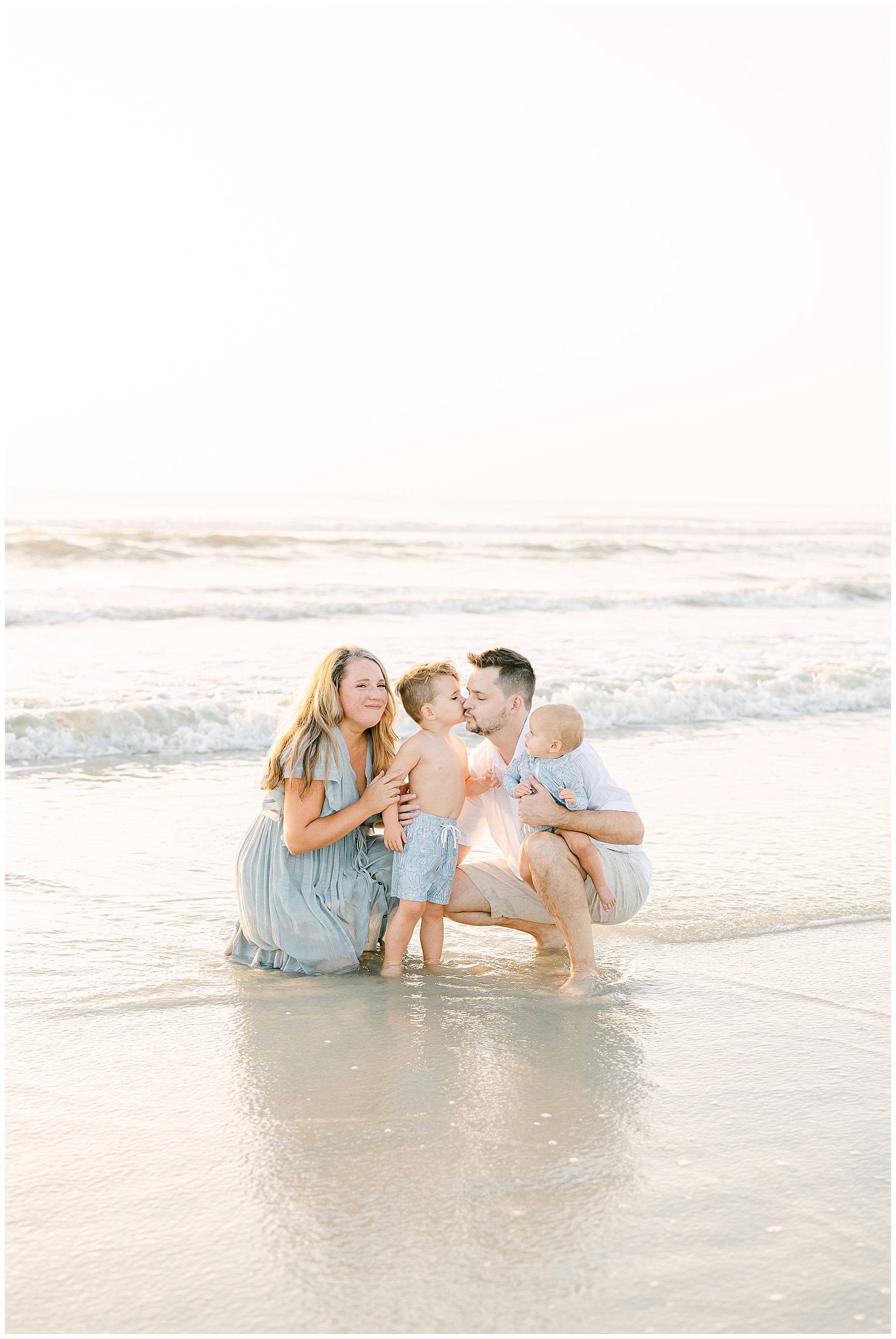 family wearing pastel colors kissing each other in the water at sunrise on the beach