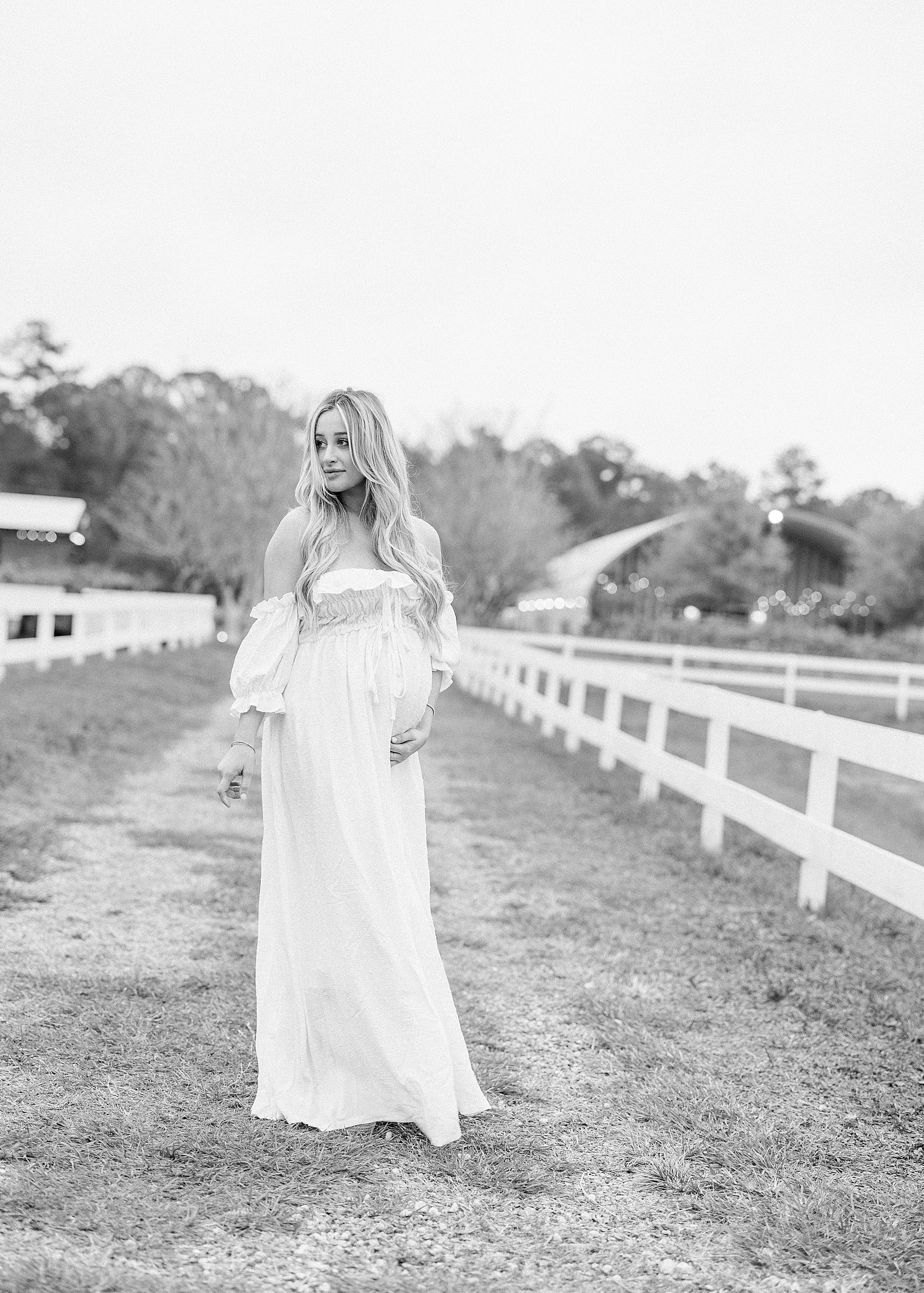 black and white maternity photo of blonde haired woman in long white dress on farm