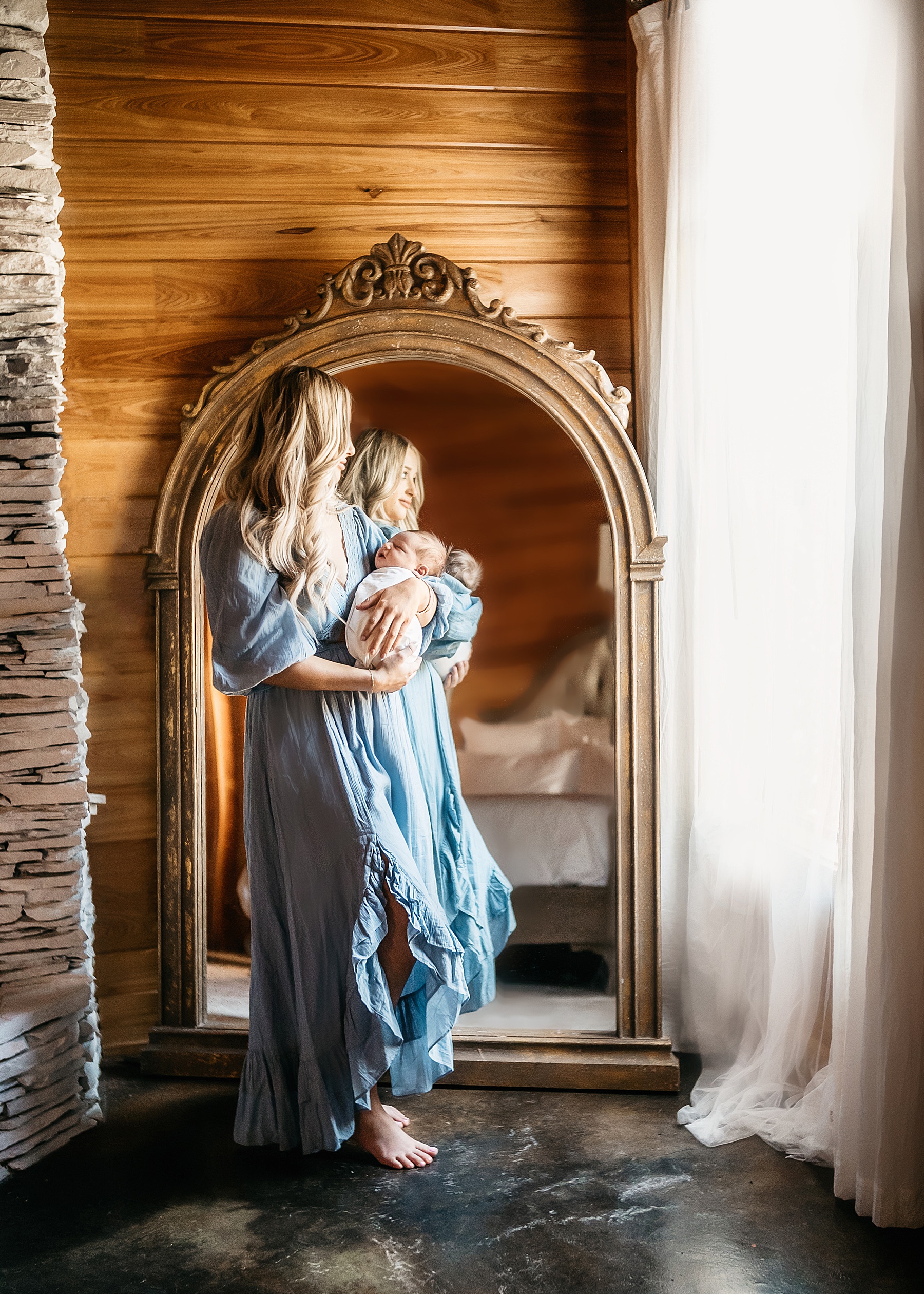 woman in blue maxi dress holding newborn baby boy wrapped in white swaddle