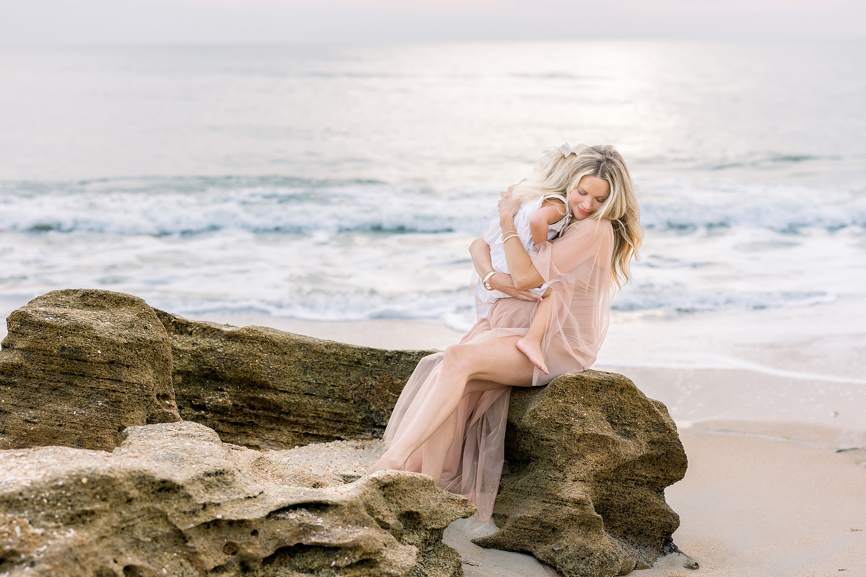 blond pregnant woman in nude dress sitting on the rocks at the beach embracing little girl in white dress